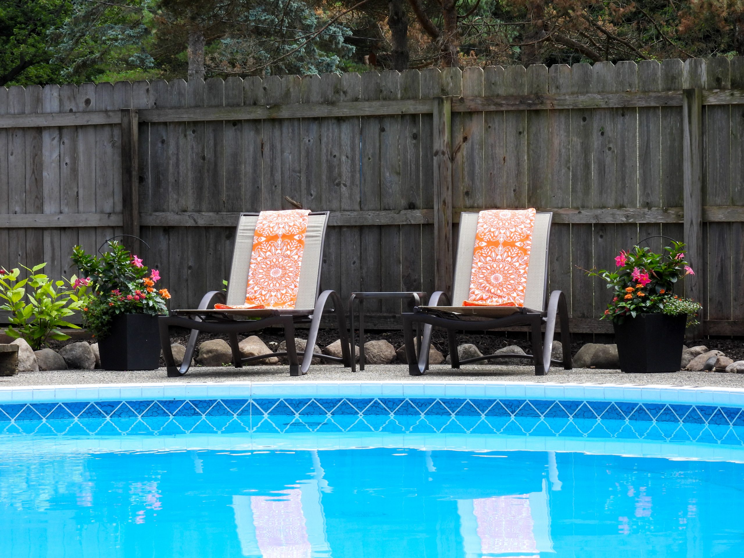 Brown fence with two outdoor lounge chairs in front of it. Chairs are next to a pool.